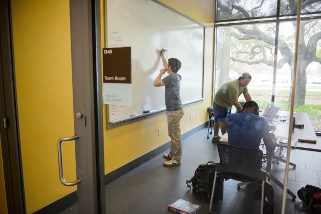 Three students working in team room, one is writing on white board. 