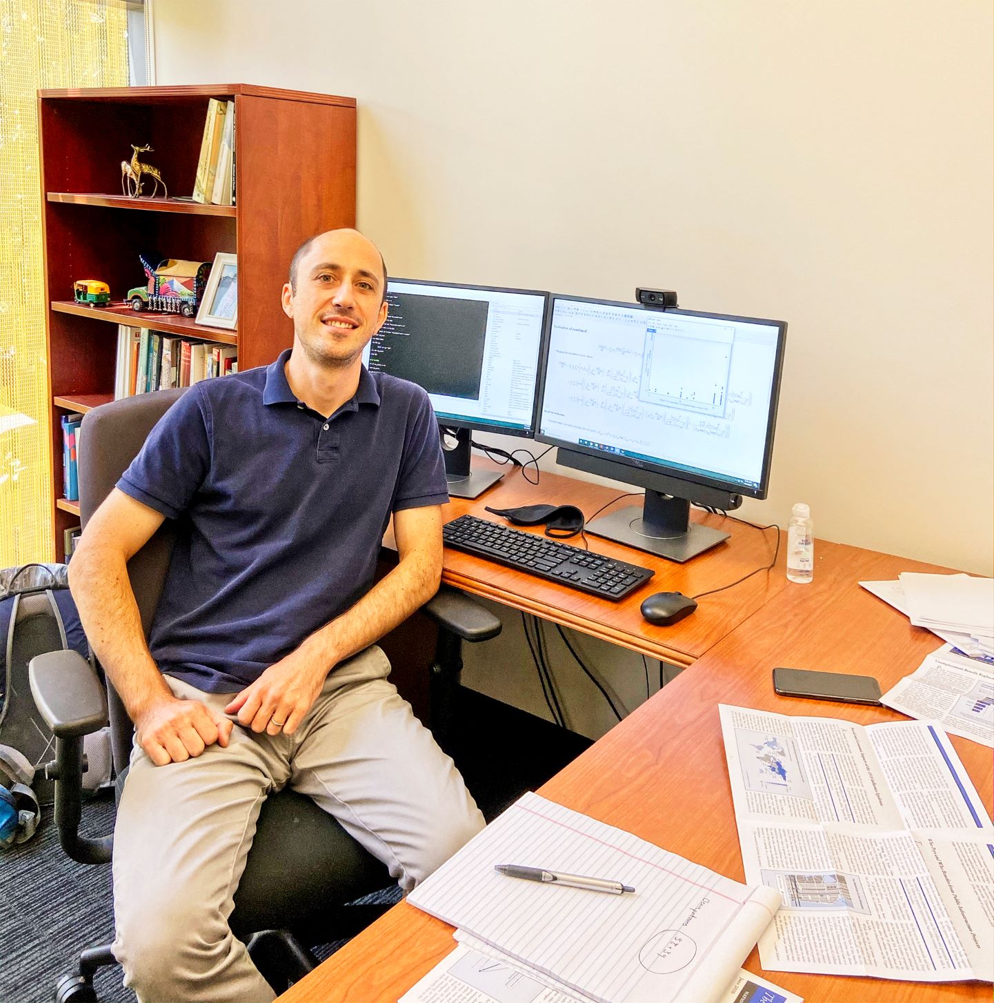 A casually-dressed Daniel Keniston leans back at his desk. His monitors show research and the surface of his desk is covered in organized paperwork.