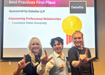 Photo of the BAP team after their regional win, each of the three team members holds his/her hand in an L as they stand before a screen announcing LSU's first place win.