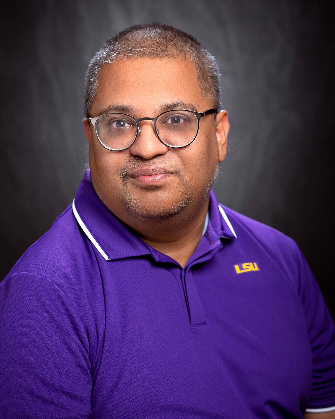a man with glasses, a purple lsu polo shirt against black photo background