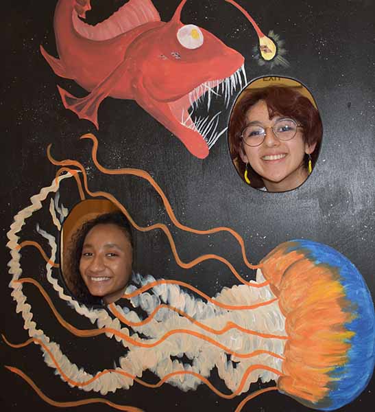 Two girls smile at the camera with a fish back drop