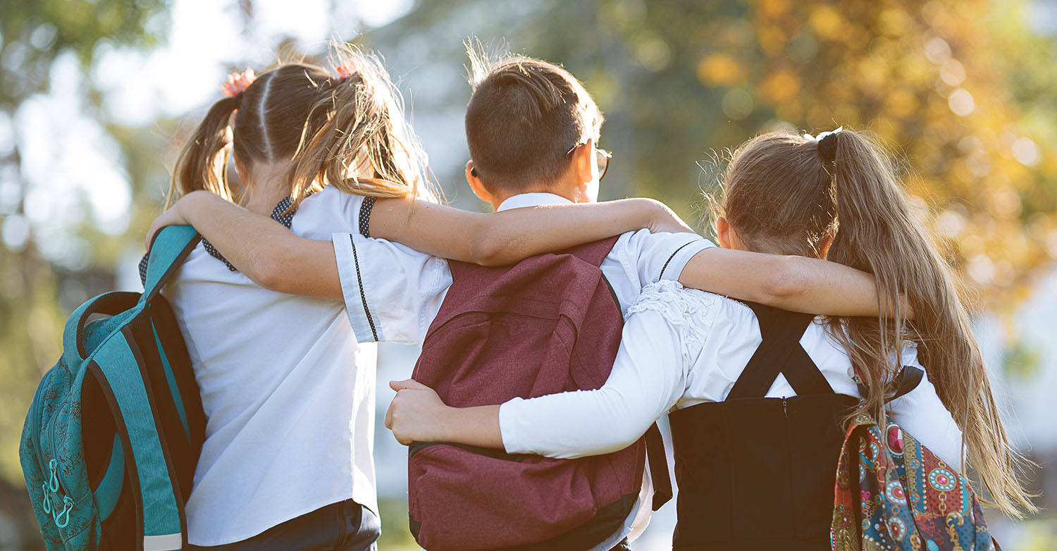 three elementary aged children wearing backpacks and their arms around each other