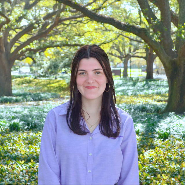 Woman in purple shirt smiling and standing in front of a tree and bushes